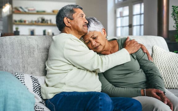 Photo of a couple embracing on a couch, looking serious, perhaps dealing with a diagnosis of Major Depressive Disorder