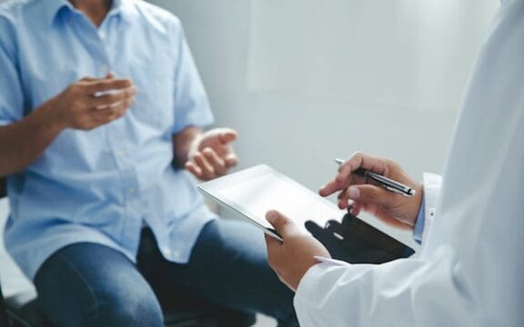 TRD Research represented by a person in a white coat holding a clipboard and talking to a patient.
